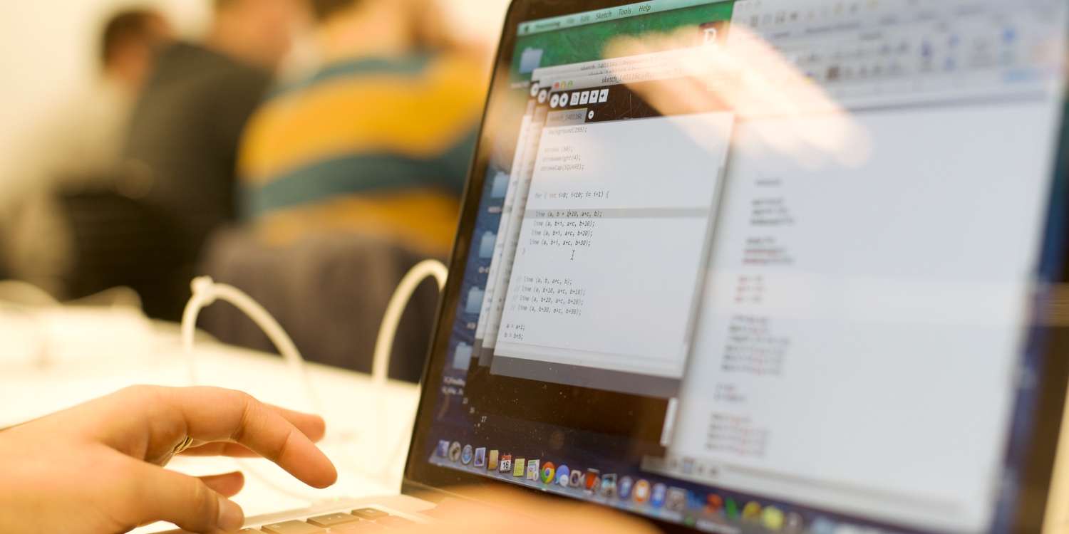 Hands at a keyboard with code on the screen and blurred people in the background